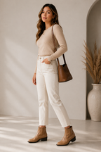 A woman stands in a bright, minimalist room wearing a beige long-sleeve top, white cropped jeans, and tan ankle boots, with a brown shoulder bag slung over her arm.